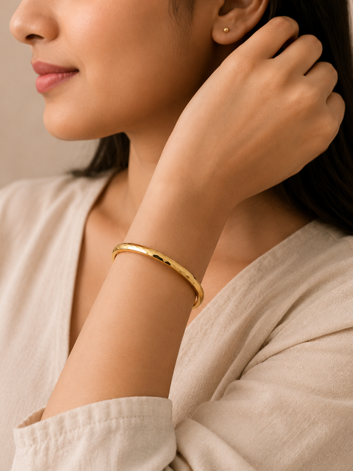 A close-up lifestyle shot of a woman wearing a polished gold bangle bracelet on her wrist, paired with a beige linen top.