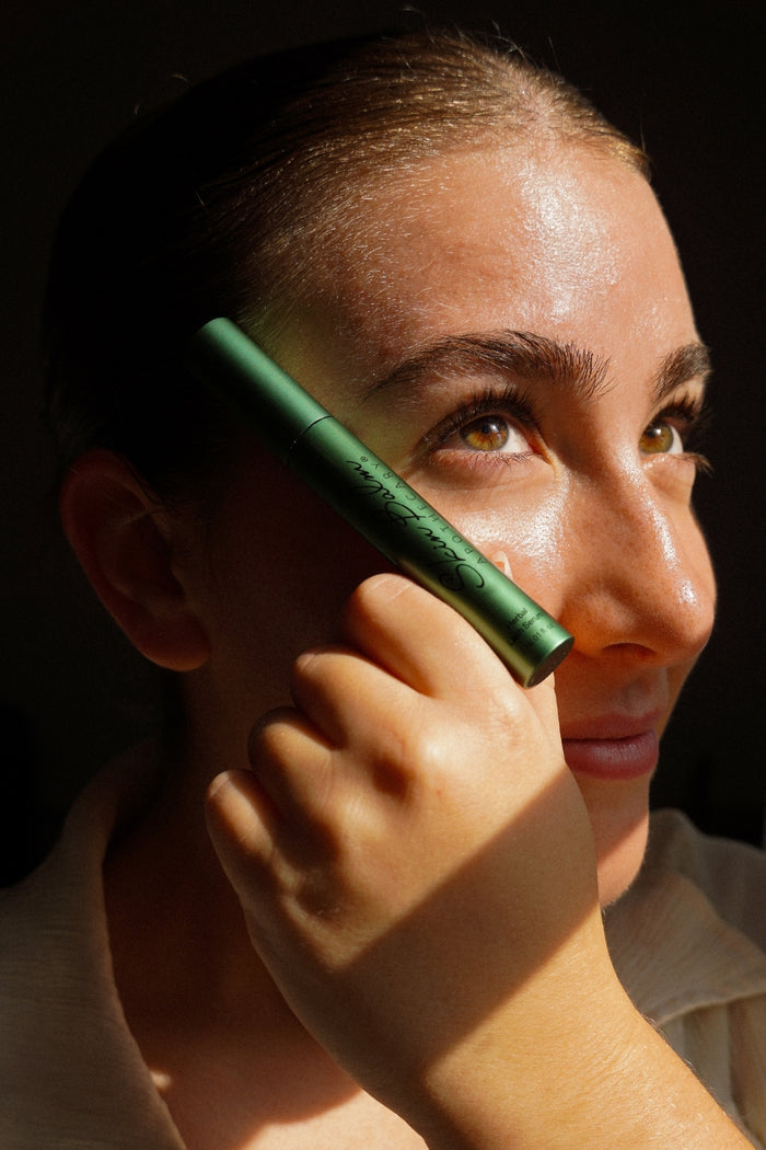 A close up shot of a woman holding the Herbal Lash Serum to her cheek against a dark, ambient background with natural lighting.