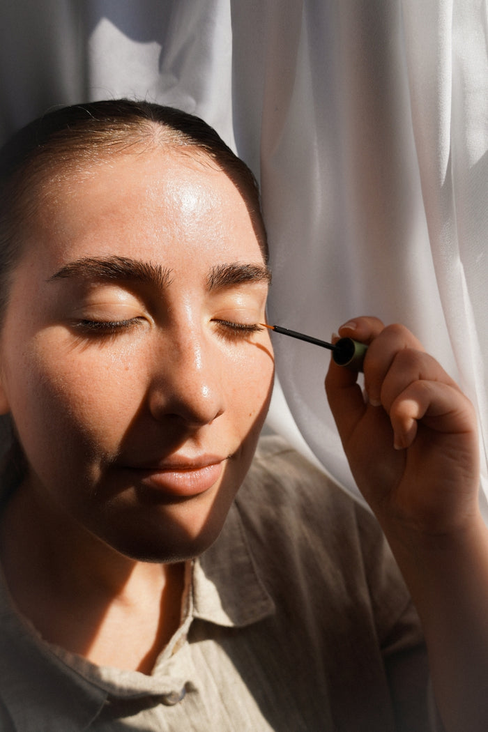 A close up shot of a woman applying the Herbal Lash Serum to her eyelash line in a dark, ambient environment; a white curtain is in the background with natural lighting.