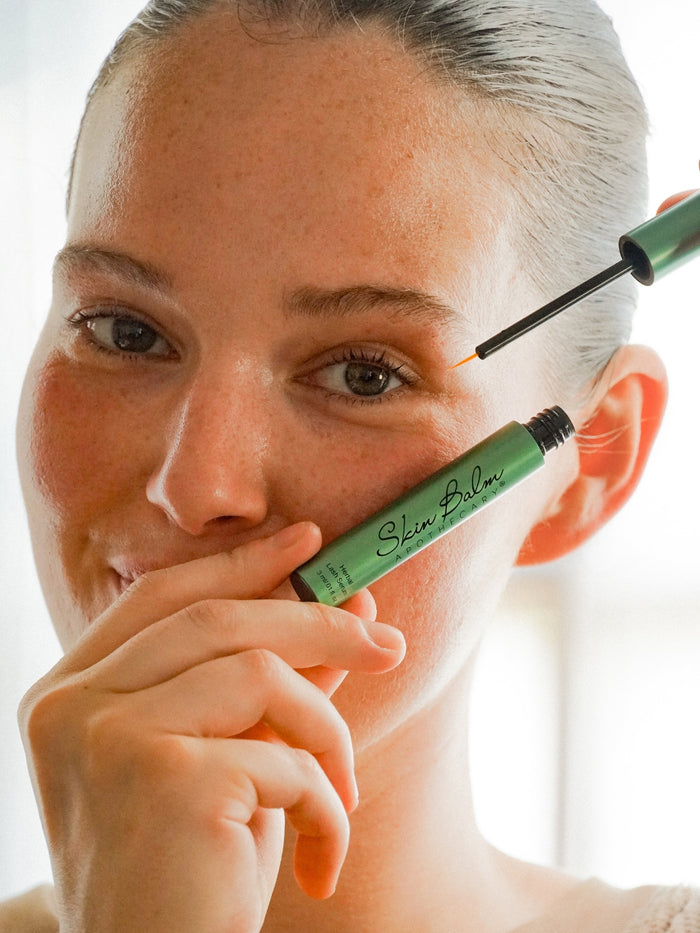 A close up shot of a woman holding the Herbal Lash Serum against her cheek with bright natural light in the background.