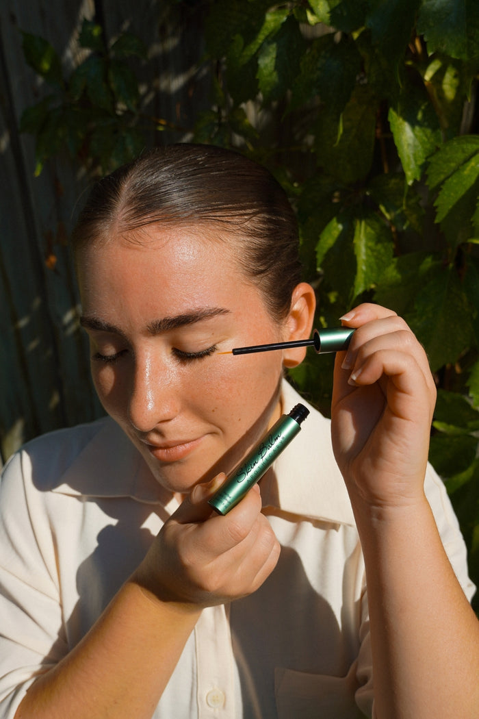 A woman applies the Herbal Lash Serum to her eyelash line outdoors with green foliage in the background.