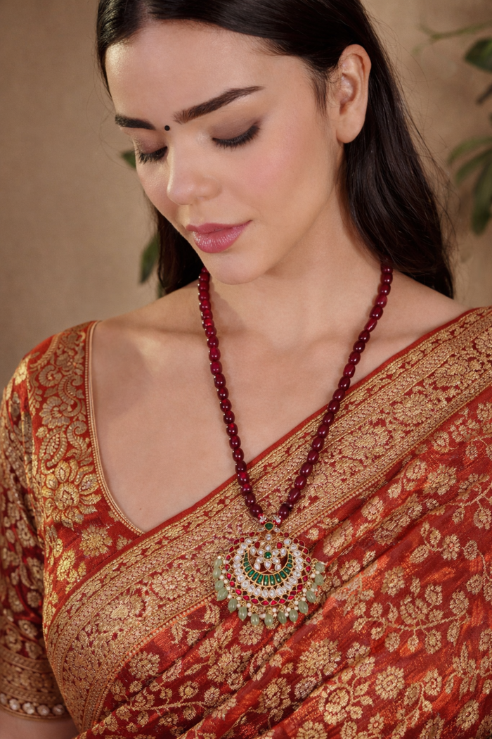A woman wearing a traditional Indian red and gold saree, featuring a prominent red beaded necklace with a large circular green and gold pendant.