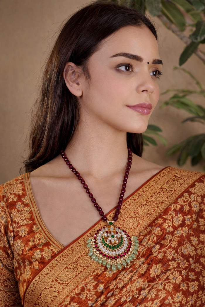 A woman wearing a traditional Indian bronze and gold Banarasi silk saree, featuring a large, ornate circular pendant necklace with maroon beads and green, white, and gold stones.